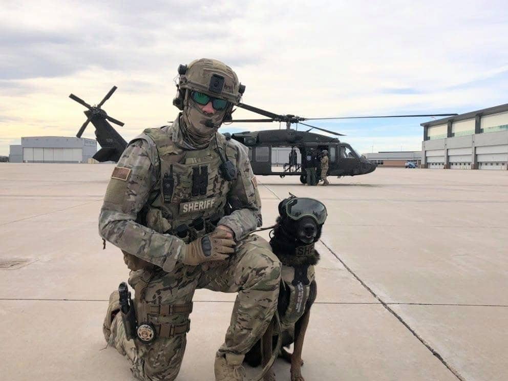 Mitch McGuinnis kneeling beside Tyr in working gear with a helicopter behind them
